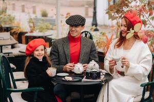 een elegant familie verzameld samen in een cafe Aan de straat. mama, pa, weinig dochter drinken thee, eten taarten. ze zijn gelukkig samen. de concept van een gelukkig familie avondeten foto