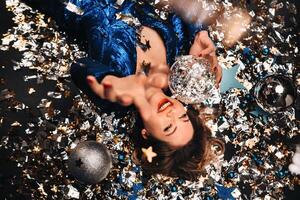 een vrouw in een blauw lovertjes jurk glimlacht en leugens Aan de verdieping onder een vallend veelkleurig confetti foto