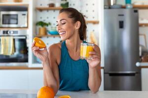 mooi jong vrouw drinken vers oranje sap in keuken. gezond eetpatroon. gelukkig jong vrouw met glas van sap en oranje Bij tafel in keuken. foto