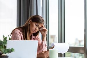 jong vrouw moe, uitgeput van werken aan het studeren moeilijk. verveeld en gefrustreerd op zoek Bij laptop. hoofd resting Aan hand. helder ruimte groot ramen. Bij huis concept. spanning concept. foto