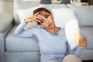 vrouw zet hoofd Aan sofa kussens voelt Sloom ten gevolge ondraaglijk warmte, golven hand- ventilator koel haarzelf, heet zomer vlak zonder airco klimaat controle systeem concept foto