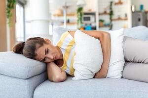 vrouw aan het liegen Aan sofa op zoek ziek in de leven kamer. mooi jong vrouw aan het liegen Aan bed en Holding handen Aan haar maag. vrouw hebben pijnlijk maagpijn Aan bed, menstruatie- periode foto