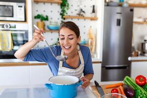 gelukkig jong vrouw Koken proeverij avondeten in een pot staand in modern keuken Bij huis. huisvrouw voorbereidingen treffen gezond voedsel glimlachen . huishouden en voeding. dieet recepten concept foto