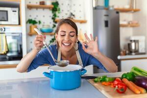 gelukkig jong vrouw Koken proeverij avondeten in een pot staand in modern keuken Bij huis. huisvrouw voorbereidingen treffen gezond voedsel glimlachen . huishouden en voeding. dieet recepten concept foto