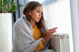 onwel vrouw huurder in deken zitten in verkoudheid leven kamer hand- Aan oud radiator.lijden van gebrek van warmte . ongezond jong vrouw worstelen van kilte bevriezen Bij huis. Nee verwarming concept. foto