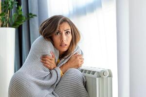 onwel vrouw huurder in deken zitten in verkoudheid leven kamer hand- Aan oud radiator.lijden van gebrek van warmte . ongezond jong vrouw worstelen van kilte bevriezen Bij huis. Nee verwarming concept. foto