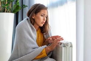 onwel vrouw huurder in deken zitten in verkoudheid leven kamer hand- Aan oud radiator.lijden van gebrek van warmte . ongezond jong vrouw worstelen van kilte bevriezen Bij huis. Nee verwarming concept. foto