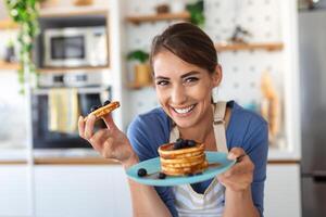 jong vrouw maken pannekoeken Bij keuken. jong huisvrouw genieten van bosbes pannekoeken voor ontbijt foto