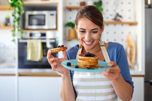 jong vrouw maken pannekoeken Bij keuken. jong huisvrouw genieten van bosbes pannekoeken voor ontbijt foto