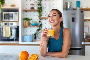mooi jong vrouw drinken vers oranje sap in keuken. gezond eetpatroon. gelukkig jong vrouw met glas van sap en oranje Bij tafel in keuken. foto