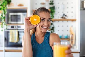 mooi jong vrouw drinken vers oranje sap in keuken. gezond eetpatroon. gelukkig jong vrouw met glas van sap en oranje Bij tafel in keuken. foto