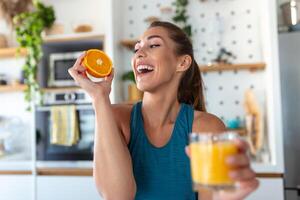 mooi jong vrouw drinken vers oranje sap in keuken. gezond eetpatroon. gelukkig jong vrouw met glas van sap en oranje Bij tafel in keuken. foto