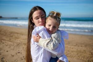 jong ma voorzichtig knuffels haar weinig kind meisje, staand met haar ogen Gesloten Aan de zanderig strand tegen de golven achtergrond foto