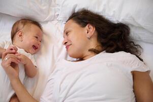 gelukkig jong moeder aan het liegen Aan de bed met haar aanbiddelijk baby jongen 4 maanden oud. mam en kind communiceren samen glimlachen foto