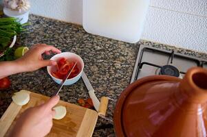 top visie van bovenstaand van een huisvrouw hakken uien en tomaten Aan een snijdend bord. een klei tajine pot Aan een staal fornuis foto