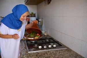 glimlachen moslim vrouw in blauw hijaab, opening deksel van een klei schotel terwijl Koken groenten in Marokkaans tajine Bij huis keuken foto