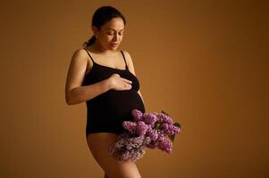 gravid toekomst moeder, zwanger vrouw in zwart bodysuit, voorzichtig strelen haar groot buik, geïsoleerd beige studio achtergrond foto