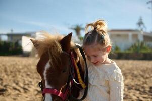 schattig blond weinig kind meisje strelen een pony, glimlachen en genieten van gelukkig weekend buitenshuis. kinderen en liefde voor dieren foto