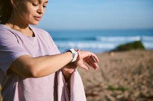 jong sportief vrouw controle pulse of hart tarief mobiel app Aan slim polshorloge, staand Aan de strand na ochtend- rennen foto