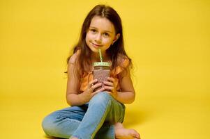 mooi kind meisje 6 jaren oud, Holding glas met heerlijk cocktail, glimlachen op zoek Bij camera, geel backdrop foto