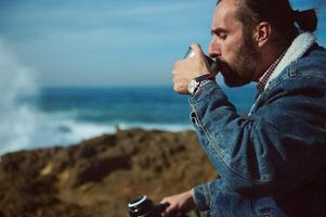 ontspannen gebaard mannetje toerist reiziger avonturier zittend Aan een rotsachtig klif door oceaan en drinken water van een thermosfles foto