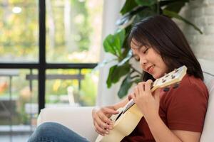 gelukkig Aziatisch vrouw zittend Aan sofa in leven kamer en beoefenen ukulele in vrije tijd. ontspanning en inspiratie van artiest. concept van muziek, kom tot rust en hobby werkzaamheid genieten in vrije tijd foto