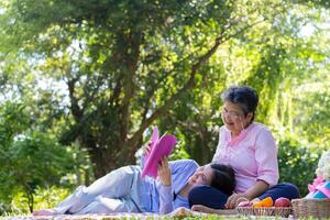 Aziatisch dochter slapen Aan moeder ronde en lezing boek Aan picknick mat in de park. een gelukkig senior vrouw praat met haar dochter. concept van gezondheidszorg en ouderen verzorger ondersteuning onderhoud. foto