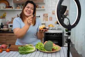 Aziatisch zwanger blogger op zoek Bij smartphone camera, ze is leven streaming Koken klasse voor zwanger. Aziatisch vrouw staand gelukkig glimlachen Bij de keuken teller voorbereidingen treffen vers biologisch salade. foto