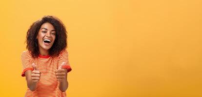 vrouw zeker weet hoe boost humeur. portret van vriendelijk ogend elegant en uitgaand Afrikaanse Amerikaans vriendin met gekruld kapsel tonen duimen omhoog in Leuk vinden en goedkeuring akkoord gaan naar vriend idee foto