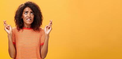 van je middel omhoog schot van hoopvol bezorgd en angstig charmant met een donkere huid vrouw met afro kapsel in modieus gestreept t-shirt bijten lip nerveus op zoek omhoog kruispunt vingers voor mooi zo geluk terwijl bidden foto