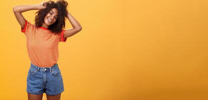 tijd begin leven leven volste. blij optimistisch vrouw hebben pret gedurende vakantie kantelen hoofd aanraken gekruld haar- en genieten van zomer zonneschijn in modieus gestreept t-shirt en shorts over- oranje muur foto