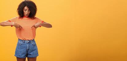 portret van bezorgd ondervraagd en verrast Afrikaanse Amerikaans vrouw met gekruld kapsel op zoek en richten Bij maag of buik gevoel ongemak en problemen met Gezondheid over- oranje achtergrond foto