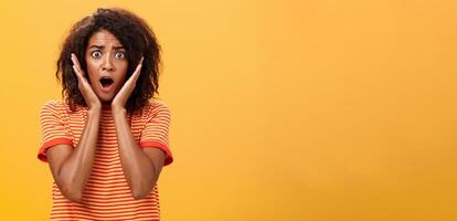 van je middel omhoog schot van geschokt bezorgd in paniek raken Afro-Amerikaans vrouw met afro kapsel in modieus t-shirt hijgen Holding palmen Aan gezicht van verrassing poseren verontrust tegen oranje muur foto