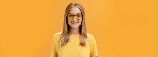elegant zelfverzekerd charmant Europese vrouw met eerlijk haar- en gebruind huid in geel t-shirt en zonnebril glimlachen breed geamuseerd en vrolijk staren Bij camera poseren tegen oranje achtergrond foto