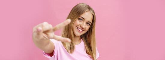 vredig en blij mooi jong meisje met Rechtdoor blond haar- in t-shirt trekken hand- met vrede teken naar camera, kantelen hoofd glimlachen vriendelijk poseren tegen roze achtergrond foto