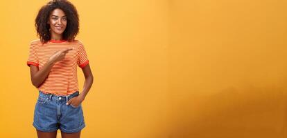 tonen perfect plaats voor reclame. charmant zorgeloos en zelfverzekerd jong elegant Afro-Amerikaans vrouw met afro kapsel Holding hand- in zak- terloops richten Rechtsaf over- oranje muur foto