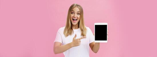 vrouw geven aanbeveling kopen digitaal tablet richten Bij apparaat scherm met inhoudsopgave vinger glimlachen enthousiast en verheugd vertellen eigen mening over apparaatje prestatie poseren verheugd over- roze muur foto