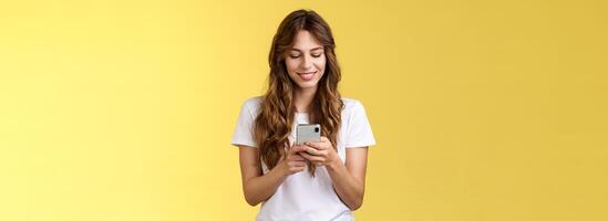 lief vrouwelijk inschrijving Kaukasisch meisje wit t-shirt jeans houden smartphone sms'en berichten vriendin glimlachen verheugd kijken teder lief glimlach mobiel telefoon scherm geel achtergrond foto