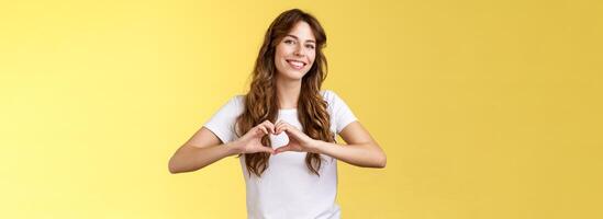 zorgeloos inschrijving aantrekkelijk vrouw gekruld lang kapsel tonen hart teken in de buurt borst uitdrukken liefde Leuk vinden zomer herinneringen glimlachen breed kantelen hoofd blij bekennen romantisch gevoelens koesteren vriendschap foto