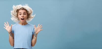 van je middel omhoog schot van onder de indruk verrast aantrekkelijk en schattig meisje met blond haar- drijvend in lucht en getatoeëerd arm jumping met verspreiding verheven palmen en geopend mond over- blauw achtergrond foto