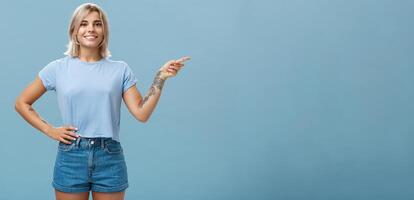 controleren het uit. portret van mooi zelfverzekerd jong Kaukasisch meisje met blond haar- richten Rechtsaf en Holding hand- Aan taille in zelfverzekerd ontspannen houding glimlachen breed over- blauw achtergrond foto