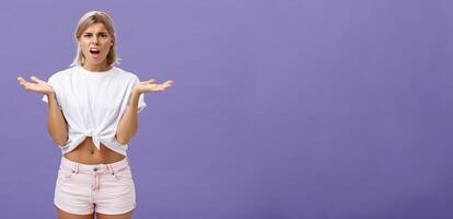 perplex en teleurstelling mooi blond vrouw leerling in wit t-shirt en roze shorts fronsen schouderophalend met verspreiding handen in de buurt schouders gezegde wat de hel over- Purper muur foto