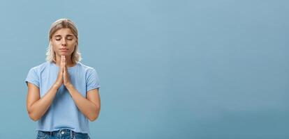 mooi soort en trouw Europese meisje met blond haar- glimlachen schattig en inschrijving Holding handen in bidden terwijl maken wens Hopelijk geloven god horen haar gebeden met Gesloten ogen over- blauw muur foto