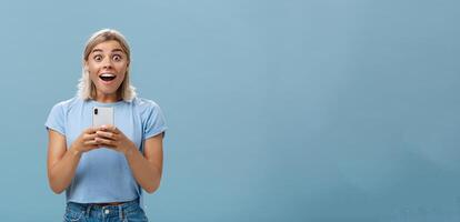 binnen- schot van versteld staan aantrekkelijk blond vrouw collega in gewoontjes t-shirt hijgen van verbazing en geluk wezen verrast staren verrast Bij camera Holding smartphone ontvangen geweldig nieuws foto