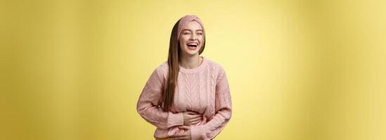 geamuseerd en vermaakt charmant jong Europese vrouw leerling lachend tot buik doet zeer van giechelen glimlachen Holding palmen Aan maag, hebben pret genieten van aan het kijken grappig komedie over- geel muur foto