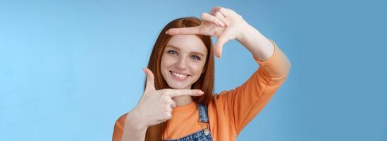 creatief mooi mooi hoor roodharige vrouw assistent zoeken inspiratie maken hand- kader kijken door verheugd glimlachen tevreden gevonden Super goed plek nemen schot staand blauw achtergrond foto