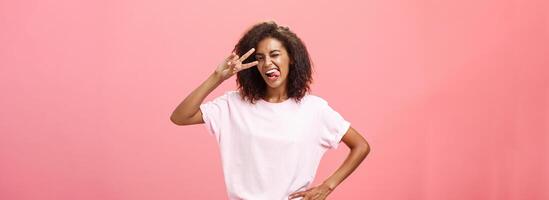 niet bang uitdrukken mezelf. blij charismatisch Afrikaanse Amerikaans vrouw in t-shirt met afro kapsel tonen tong speels en gedurfd maken vrede teken over- oog en knipogen poseren over- roze achtergrond foto