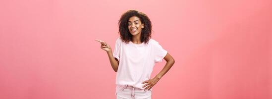 Hallo Look. portret van schattig kilte en vriendelijk elegant Afrikaanse Amerikaans vrouw met afro kapsel Holding hand- Aan taille richten Rechtsaf en glimlachen blij over- roze achtergrond foto