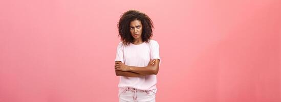 portret van beledigd somber verdrietig Afrikaanse Amerikaans vrouw vriend kruispunt armen Aan borst in bescherming gebaar fronsen op zoek van onder voorhoofd met belediging op zoek jaloers en boos over- roze muur foto