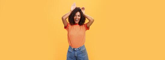 laat me worden uw konijn. portret van charmant enthousiast en charismatisch gelukkig met een donkere huid vrouw met afro kapsel Holding palmen Aan hoofd Leuk vinden dier oren maken schattig gezicht over- oranje achtergrond foto